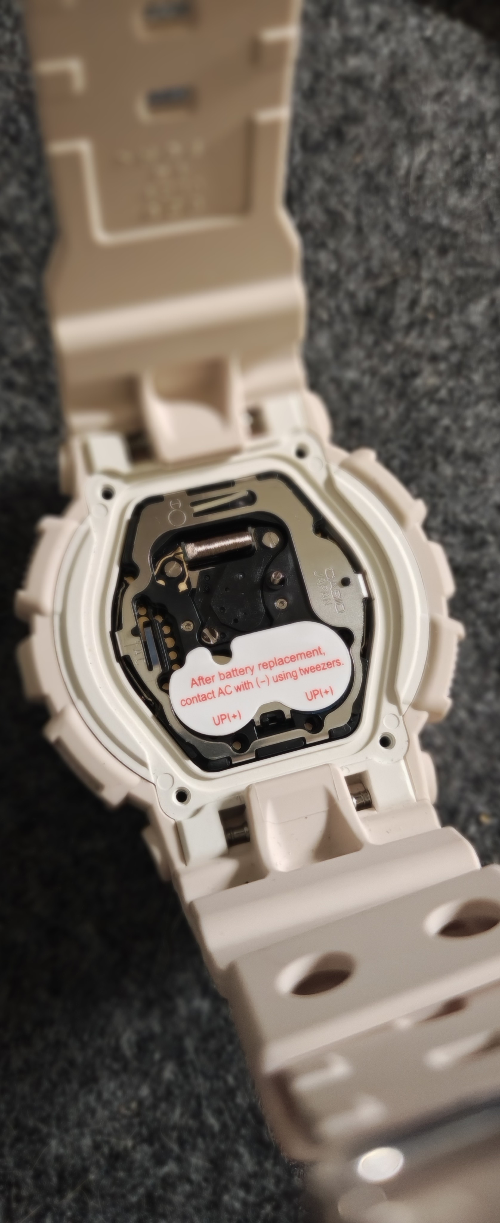 一块浅粉色手表的背面特写，金属后盖已被移除，露出内部的黑色机芯。机芯上贴有一张白色绝缘贴纸，上面印有红色的英文电池更换说明：“After battery replacement, contact AC with (-) using tweezers”（更换电池后，请使用镊子连接 AC 端和负极）。