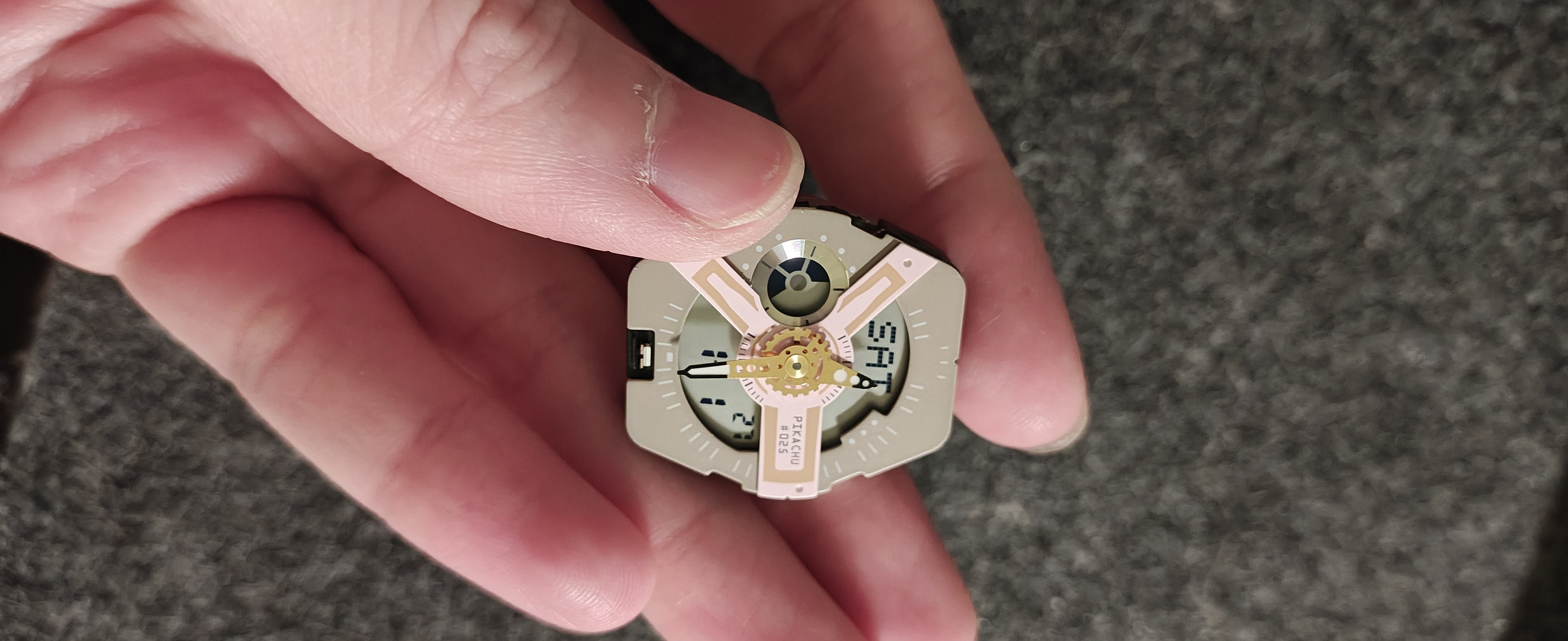 一只手拿着一个拆卸下来的圆形手表机芯。表盘为米色，带有粉色的几何闪电图案装饰和金色指针。表盘中央印有“PIKACHU #025”字样，数字显示屏显示“SAT”，表明这是一款宝可梦皮卡丘联名款手表。