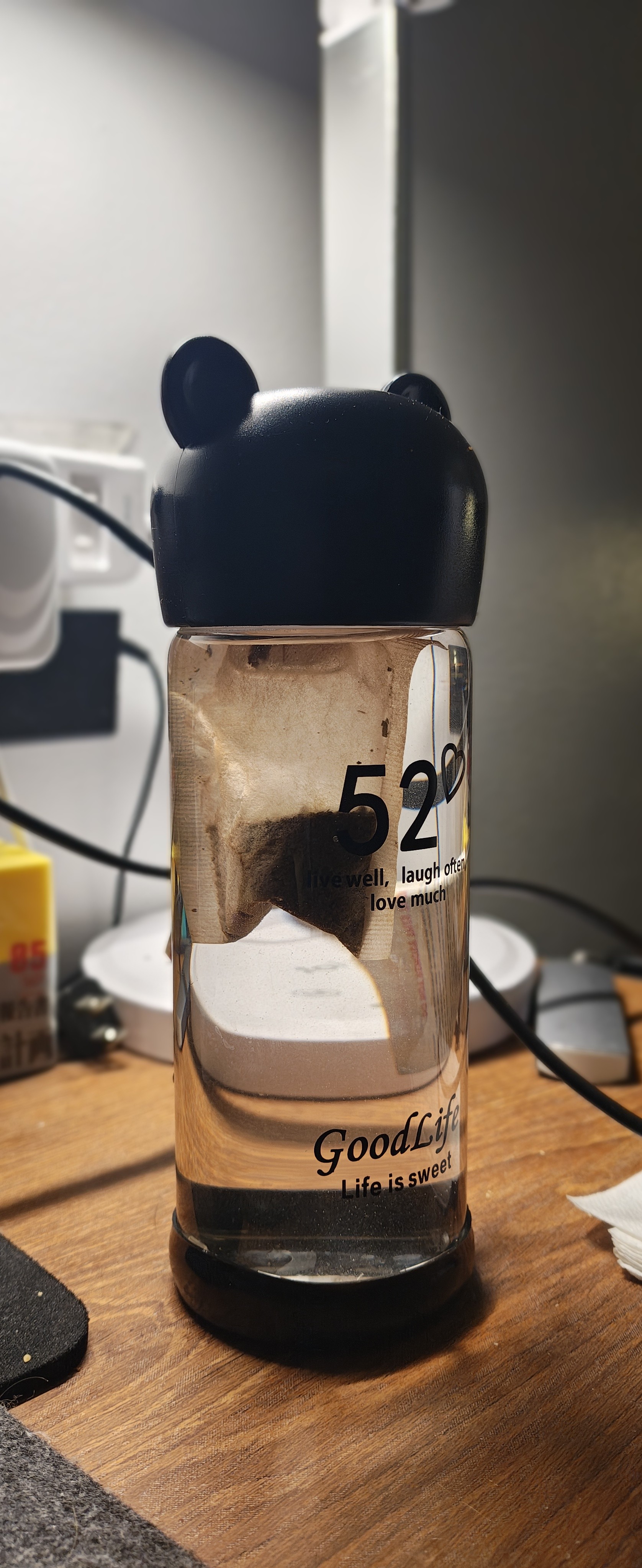 一个透明的玻璃水杯，杯身印有黑色数字“52”和文字“livewell, laugh often, love much”以及“GoodLife Life is sweet”。杯子里有一个泡在水里的茶包，杯盖是黑色的，带有两个小耳朵。