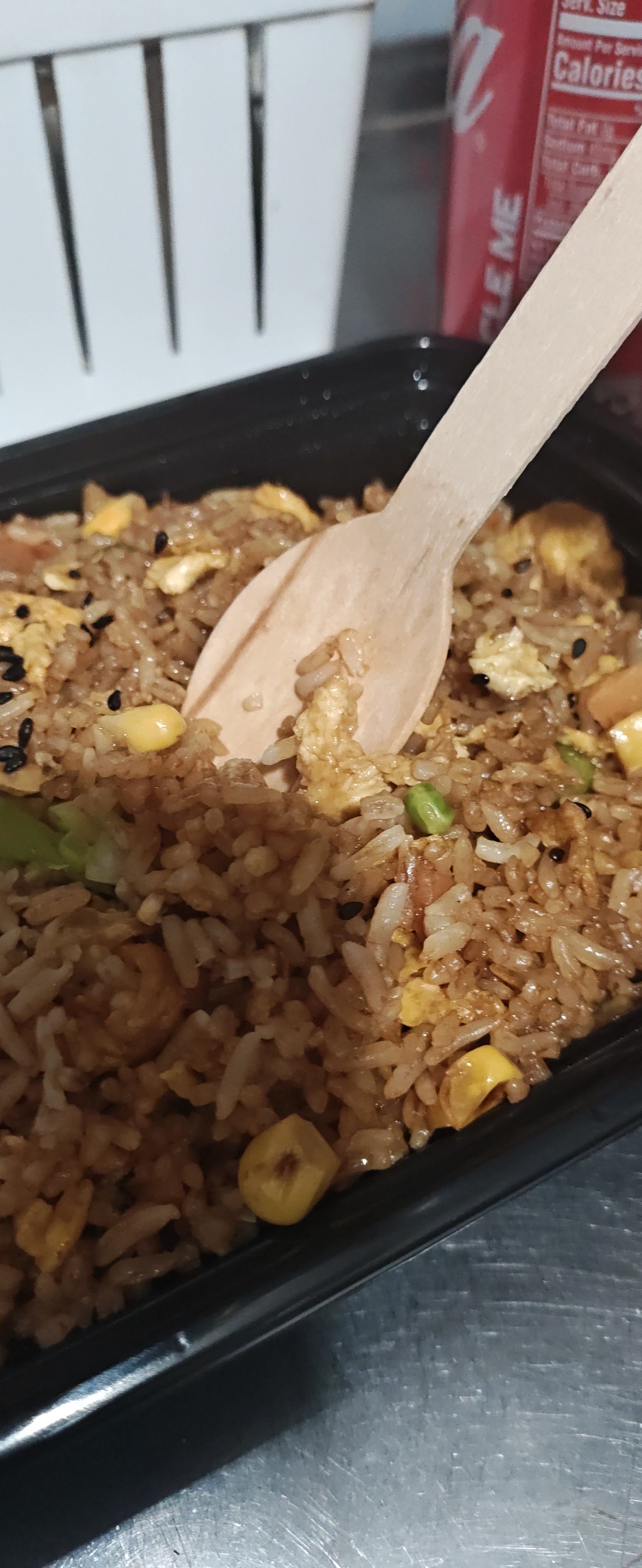 一份装在黑色外卖盒里的炒饭特写，米饭中可见鸡蛋、玉米粒、青豆和黑芝麻，一把木质勺子插在炒饭中，背景模糊处有一罐红色汽水和白色容器。