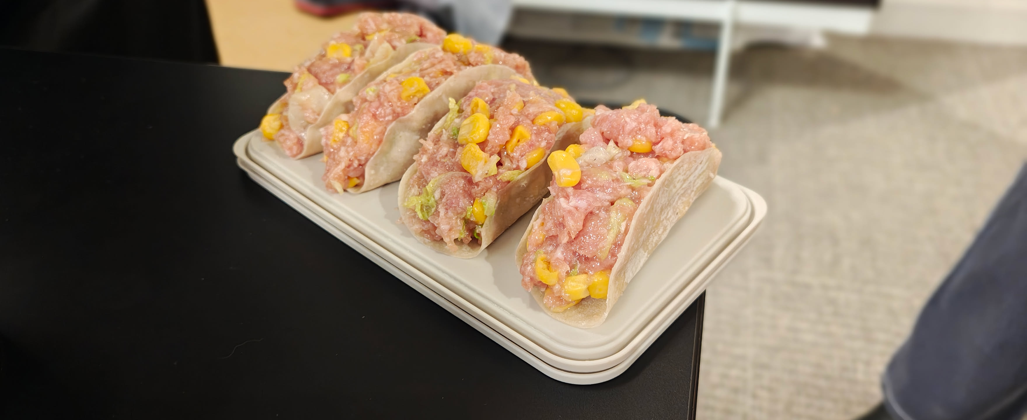 一盘模仿塔可造型的创意饺子，饺子皮像塔可壳一样立起，内填猪肉馅和玉米粒，摆放在白色便当盒上。