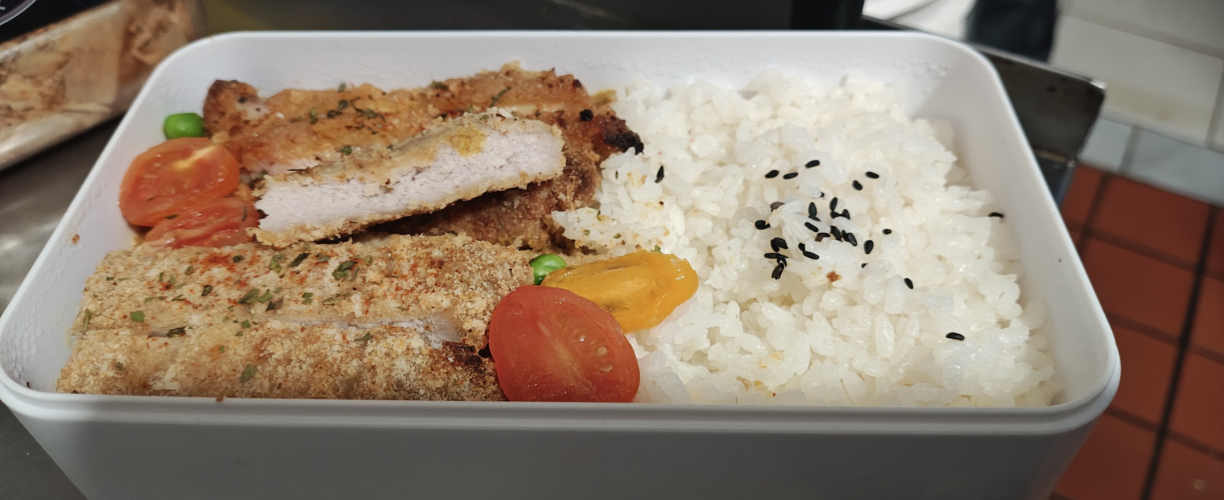 一个白色的长方形便当盒，里面装有撒了黑芝麻的白米饭、切成条状的金黄炸鸡排（可以看到内部白色的肉质），并配有红黄两色的切片圣女果和少许青豆。