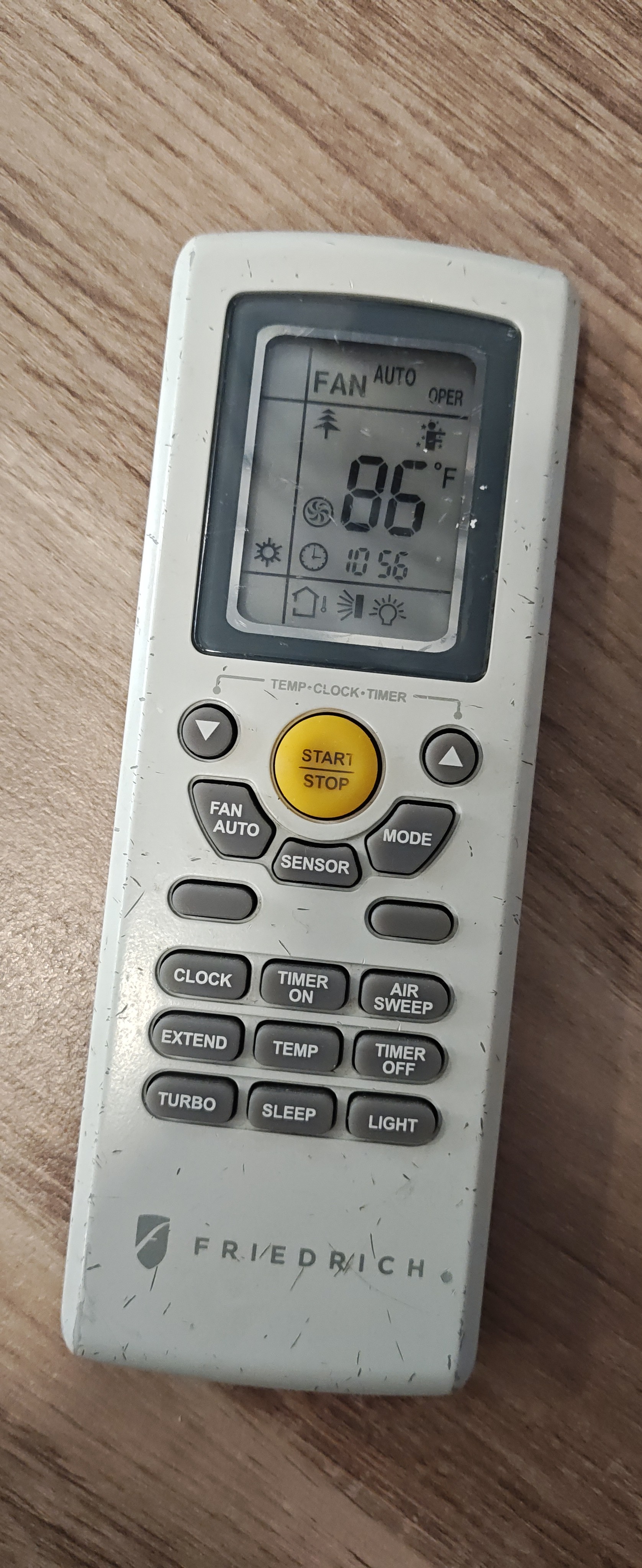 这是一张放置在浅色木纹表面上的Friedrich品牌空调遥控器的特写照片。遥控器通体呈灰白色，机身正面及边缘分布着细微的黑色划痕和磨损痕迹，显示出该设备已被长期使用。遥控器上方的LCD显示屏处于开启状态，屏幕中央醒目地显示设定温度为86华氏度（°F）。屏幕左侧的太阳图标表明空调当前设定为制热模式，顶部文字显示“FAN AUTO”，意为自动风速。屏幕中央的时钟显示时间为10:56。与另一张照片相比，该屏幕上额外显示了一个松树形状的图标（通常代表健康或换气模式）以及一个圆形的风扇图标。遥控器中部是一个醒目的黄色圆形“START/STOP”电源键，周围环绕着灰色的功能按键，底部印有深灰色的“FRIEDRICH”品牌标识。