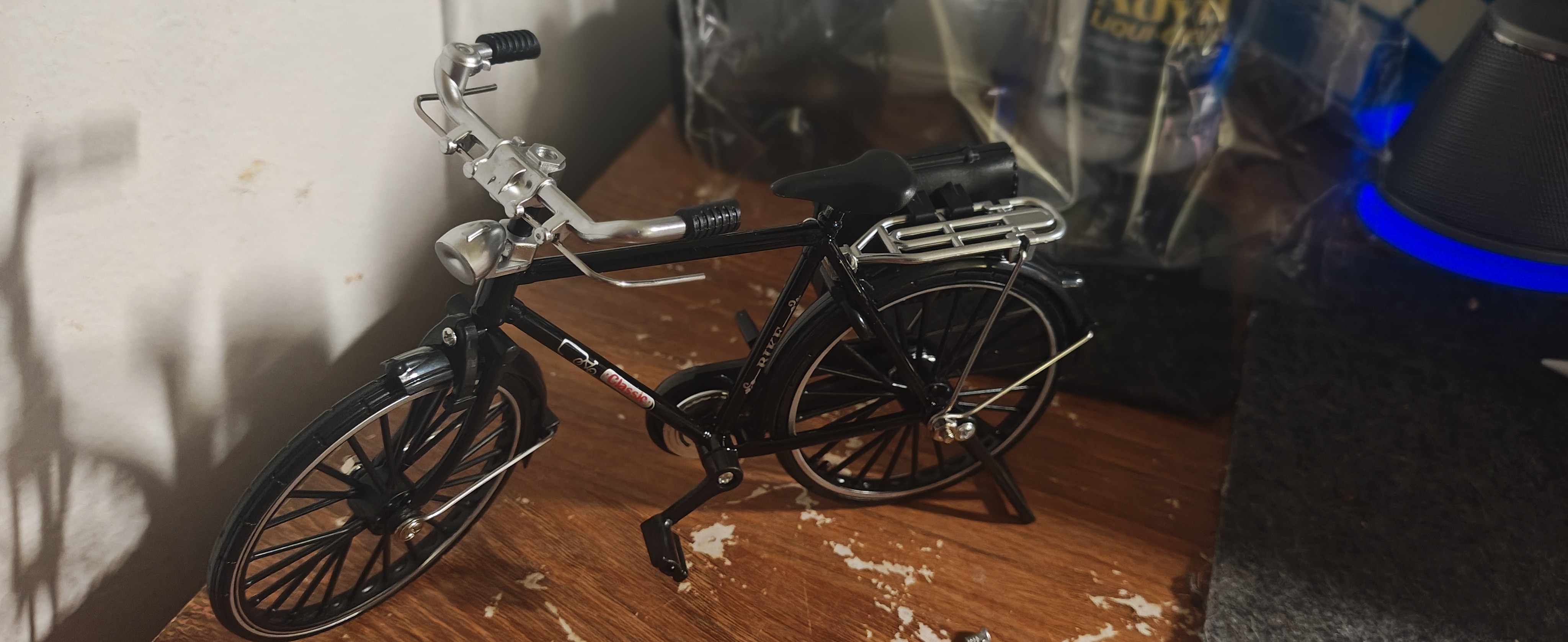 一个放置在木质表面上的黑色复古自行车微缩模型。模型细节精致，具有银色的弯曲车把、安装在前叉上的车灯、黑色的车座以及银色的后货架。自行车通过脚撑直立停放，背景略显模糊，可见部分杂物和蓝色的光映。