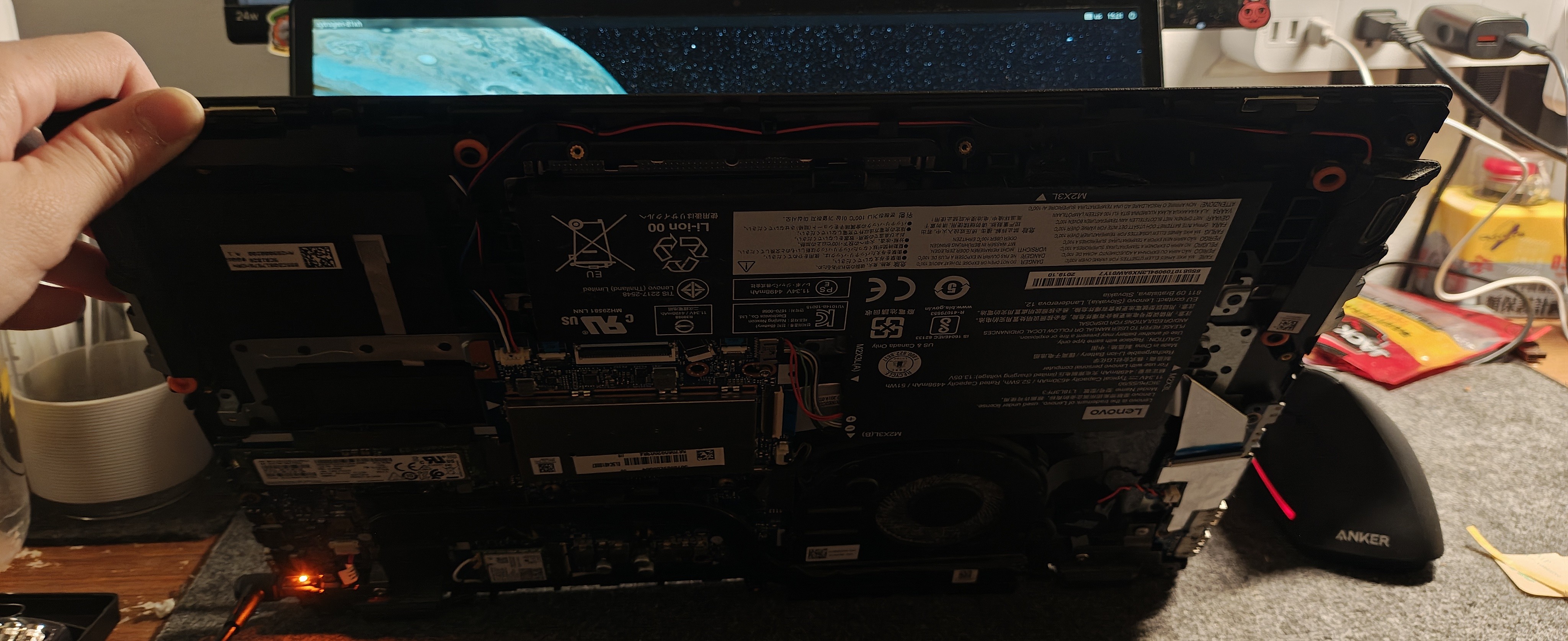 一台处于拆解维修状态的笔记本电脑的底部视角。我的左手抬起了已经移除后盖的机身，清晰地暴露了内部的硬件布局，包括占据很大面积的黑色锂电池组、绿色的主板电路、散热风扇以及连接各个组件的排线。在机身左下角可以看到一颗橙色的指示灯正在亮起。背景是略显杂乱的深灰色工作台垫，上面放着电源插座、鼠标以及其他维修设备。