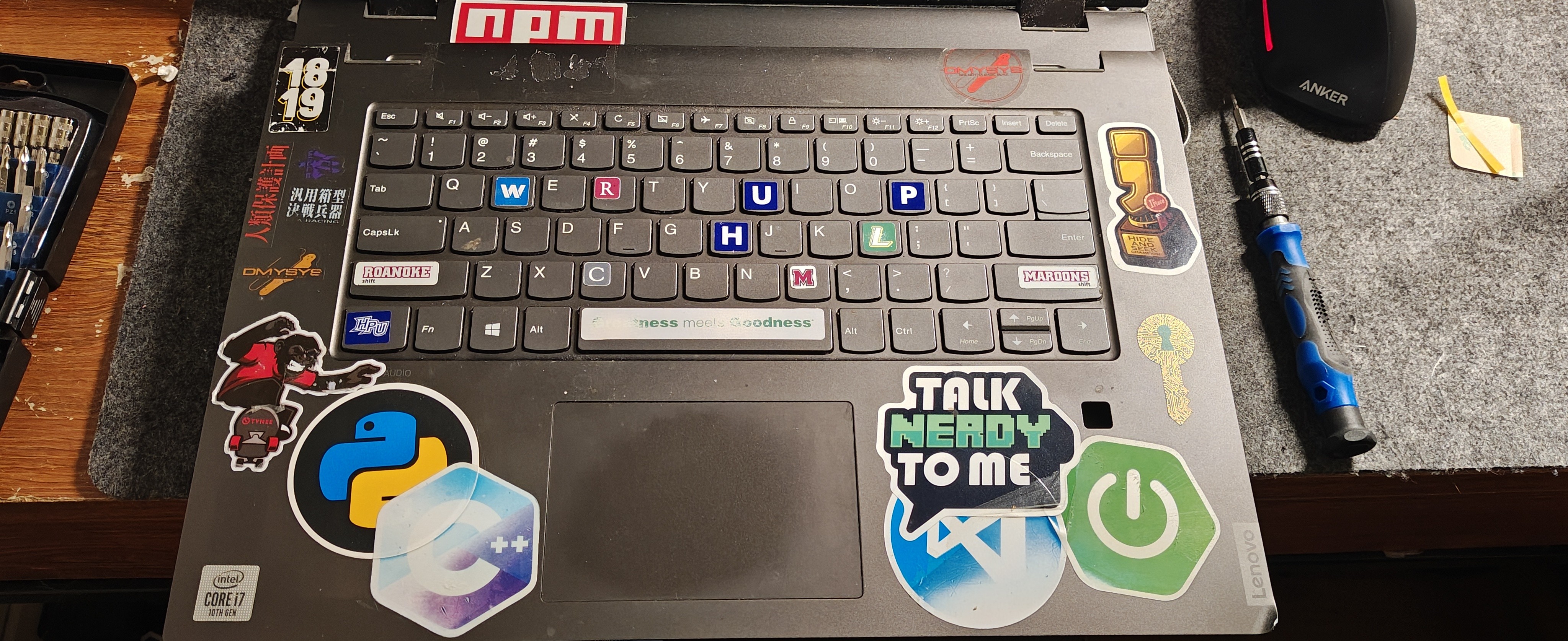 以俯视角度拍摄了一台贴满各色编程技术贴纸（如 Python、C++、Spring、npm 等）的联想笔记本电脑的 C 面。有一个被拆卸下来的旧键盘直接覆盖放置在已经安装好新键盘的机身之上。旧键盘上的部分键帽（如 WASD 和 HJKL 区域）被替换成了醒目的彩色键帽。电脑的右侧放置着一把蓝黑相间的精密螺丝刀，左侧可见一盒螺丝批头。