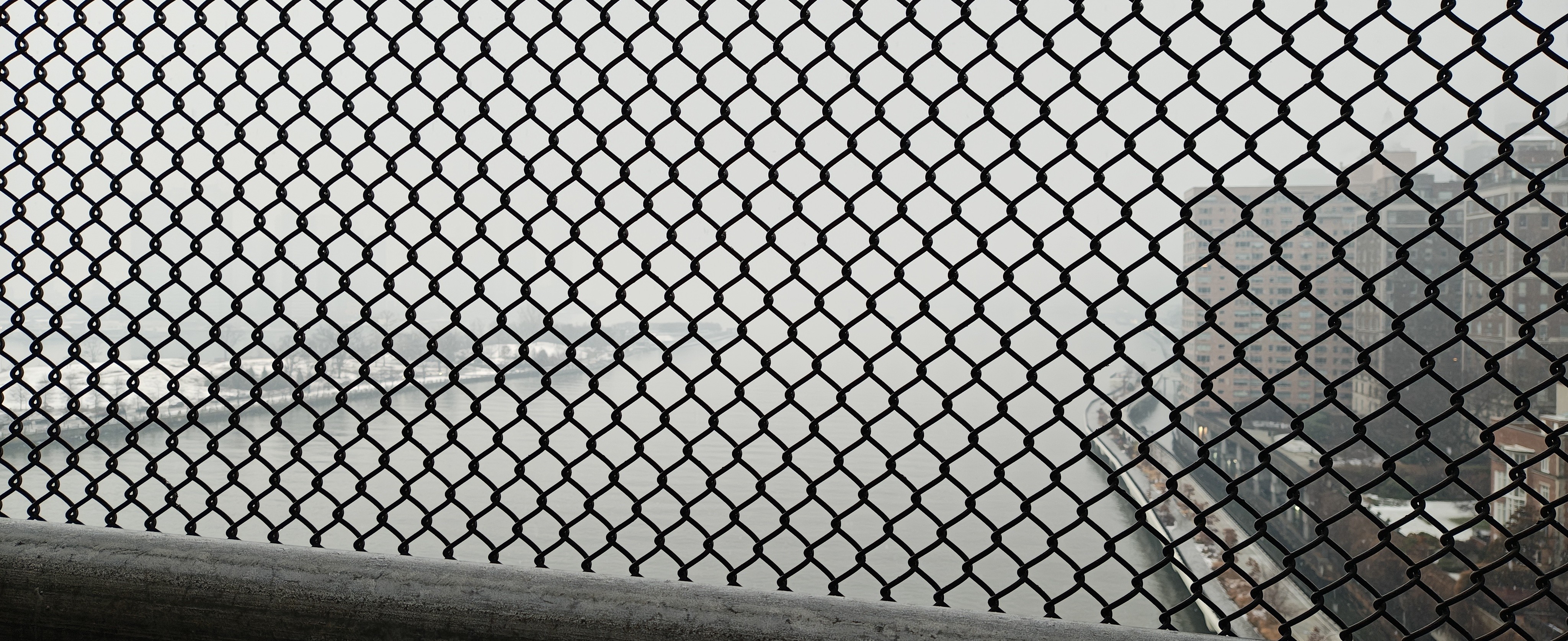 一个从高处俯瞰的城市景观，前景被密集的黑色铁丝网围栏完全遮挡。围栏的菱形网格结构占据了画面的大部分，焦点落在金属网线上，使得背景中的城市景色呈现出模糊的虚化效果。
​透过围栏可以看到，背景是一条宽阔的海湾，水面呈现深灰色。右侧排列着多栋高层建筑，建筑风格较为传统且密集。地面上覆盖着一层薄薄的积雪或霜，显示出寒冷的气候特征。整个画面色彩以灰色、黑色和暗棕色为主，天空阴沉，营造出一种冷峻、压抑且带有工业质感的氛围。