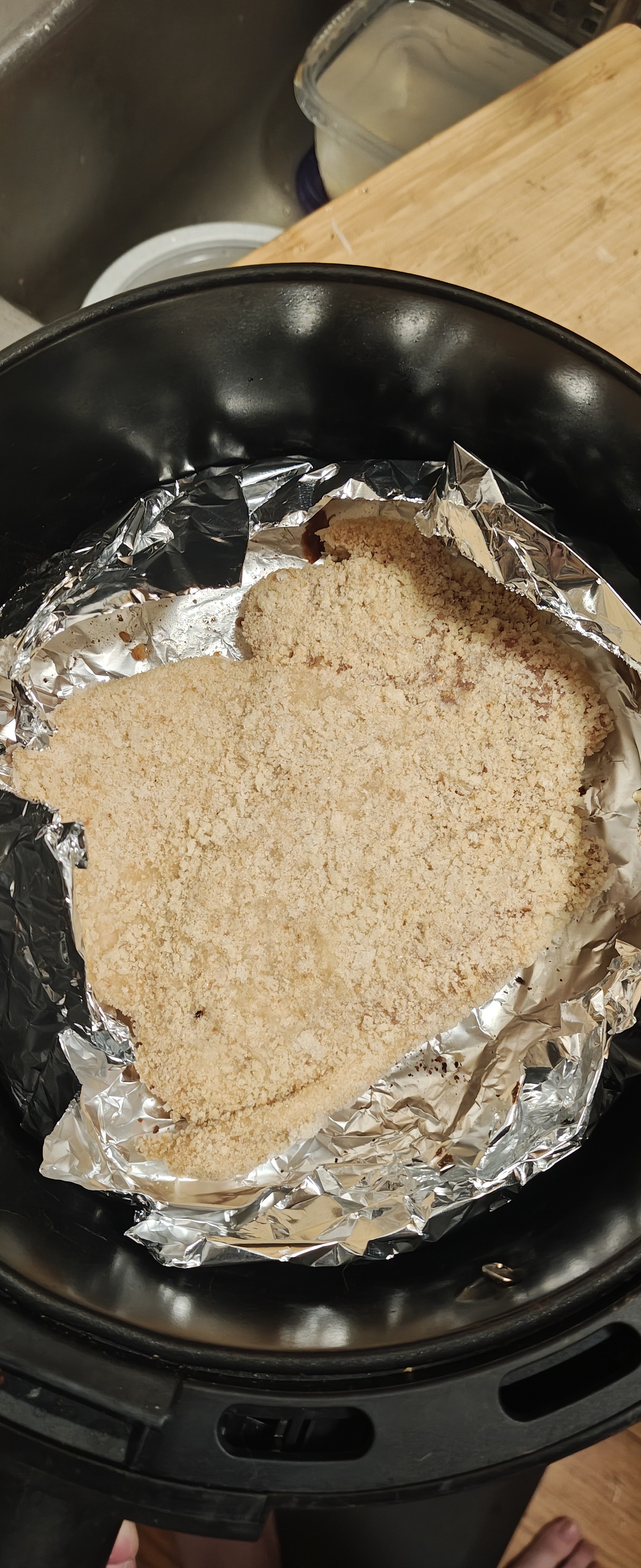 一个黑色炸篮底部铺着一层铝箔纸，上面平铺着一大块裹满面包糠的鸡腿排。
​鸡肉呈现出浅褐色的干燥粉末质感，显示出尚未开始烹饪或处于加热初期的状态。铝箔纸边缘略微卷起，紧贴炸篮内壁，整个构图从俯视角度居中拍摄，光线均匀，背景可见厨房台面的局部细节。