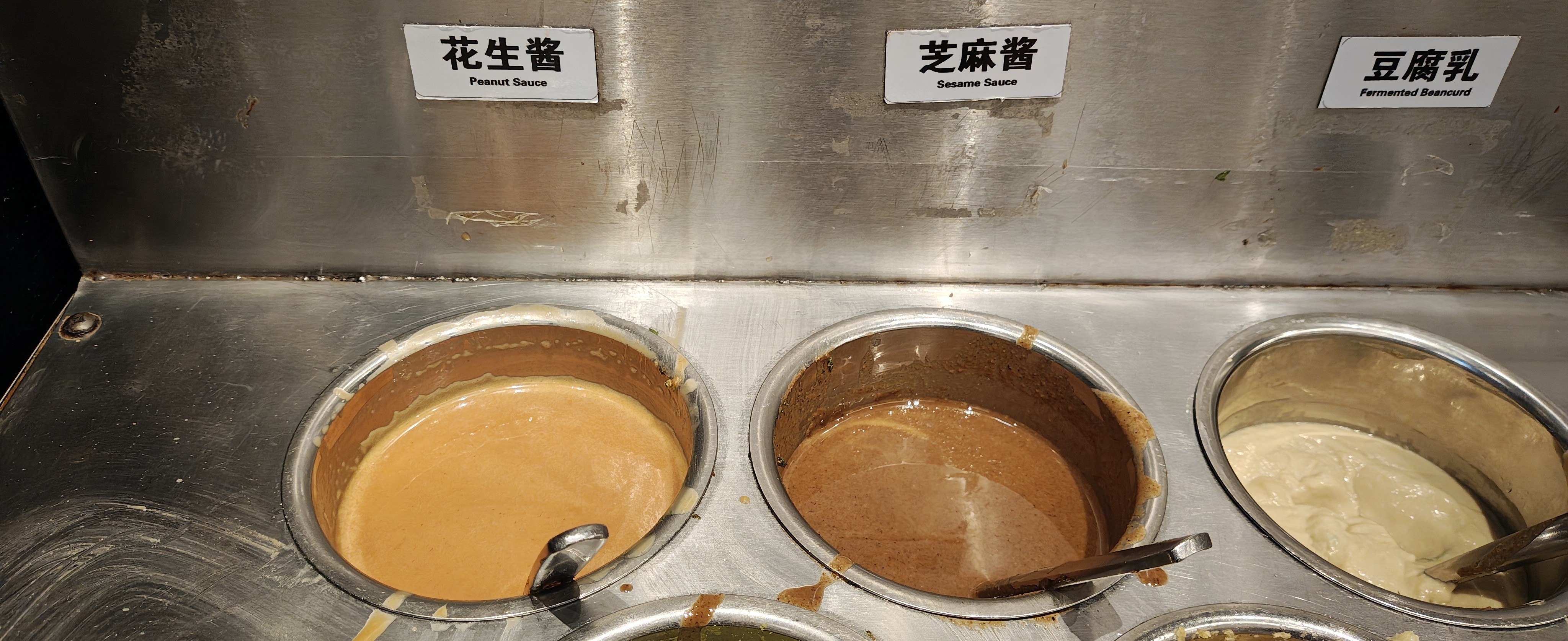 一张特写照片，展示了餐厅自助调料台上的三个不锈钢酱料桶。从左到右依次盛放着浅黄色的花生酱、深褐色的芝麻酱和白色的豆腐乳。每个酱料桶上方都贴有白底黑字的中英文标签，分别标注为「花生酱 Peanut Sauce」、「芝麻酱 Sesame Sauce」和「豆腐乳 Fermented Beancurd」。  