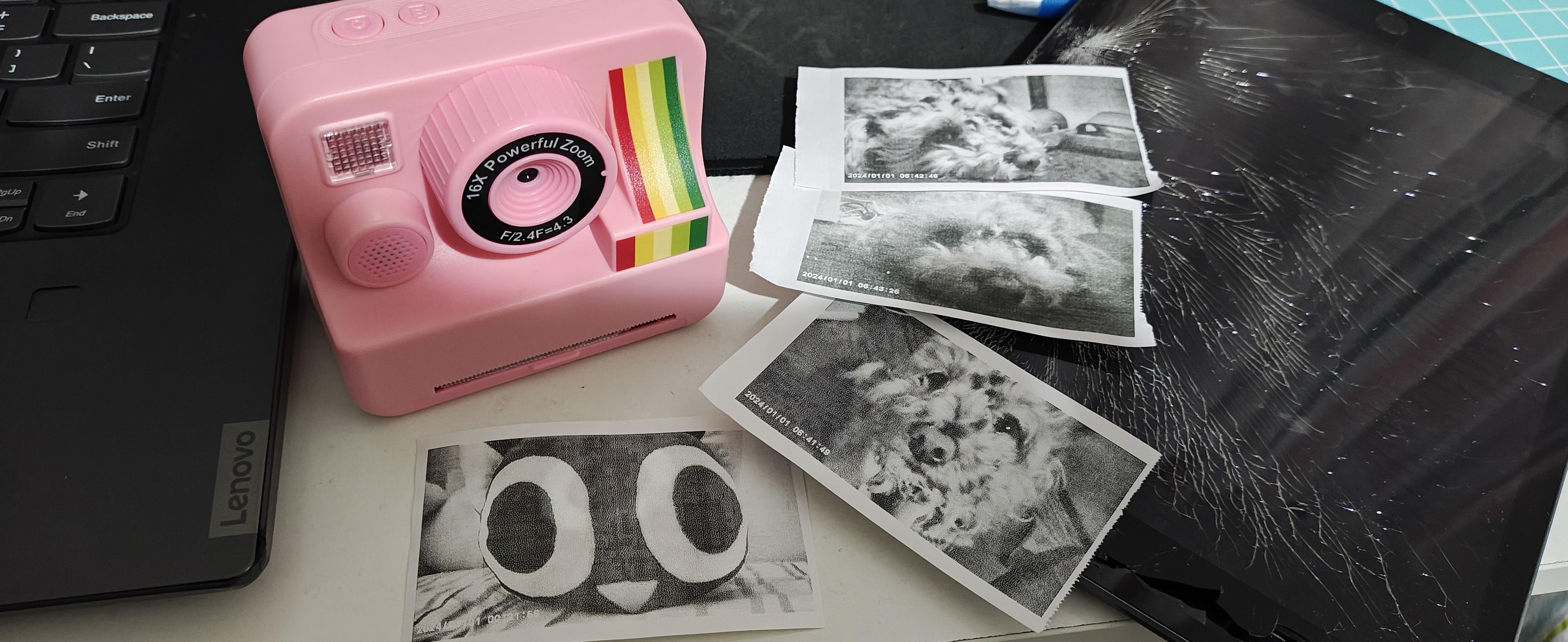 白色桌面上放置着一台粉红色的儿童热敏打印相机，旁边散落着几张印有迷你泰迪犬和罗小黑玩偶的黑白小照片；右侧是一台屏幕严重碎裂的 iPad，左侧可见黑色联想笔记本电脑的一角。