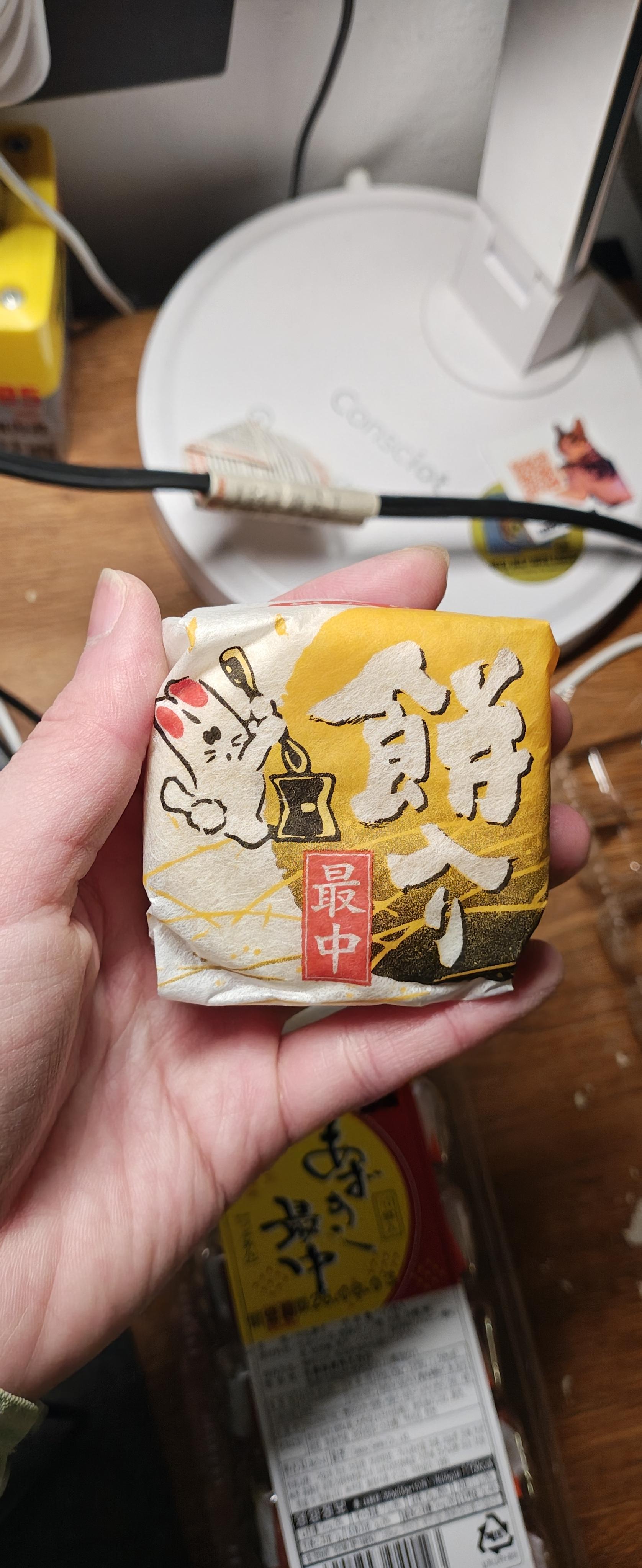 一只手握着一个方形包装的日式点心。包装纸呈现淡黄色，质感类似和纸，上面印有醒目的黑色书法字体「饼入り」（含年糕）以及红色方框内的白色文字「最中」（Monaka，一种夹心糯米皮点心）。包装左侧绘有一只可爱的白色兔子卡通形象，兔子正提着一只灯笼。背景中可以看到一个白色的圆形台灯底座以及其他未拆封的同款点心包装。